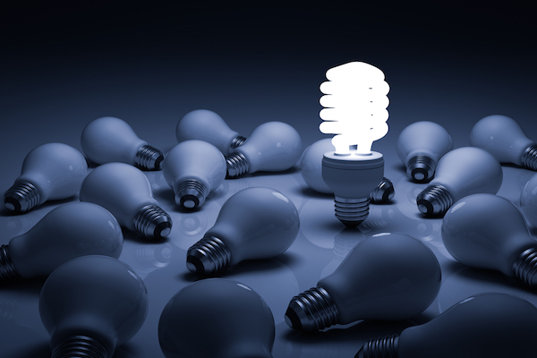 Compact Fluorescent Light Bulbs standing and shining in a dark room full of unlit, fallen conventional light bulbs