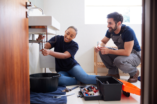 A plumber's apprentice works on the pipes beneath a sink while a licensed plumber supervises.